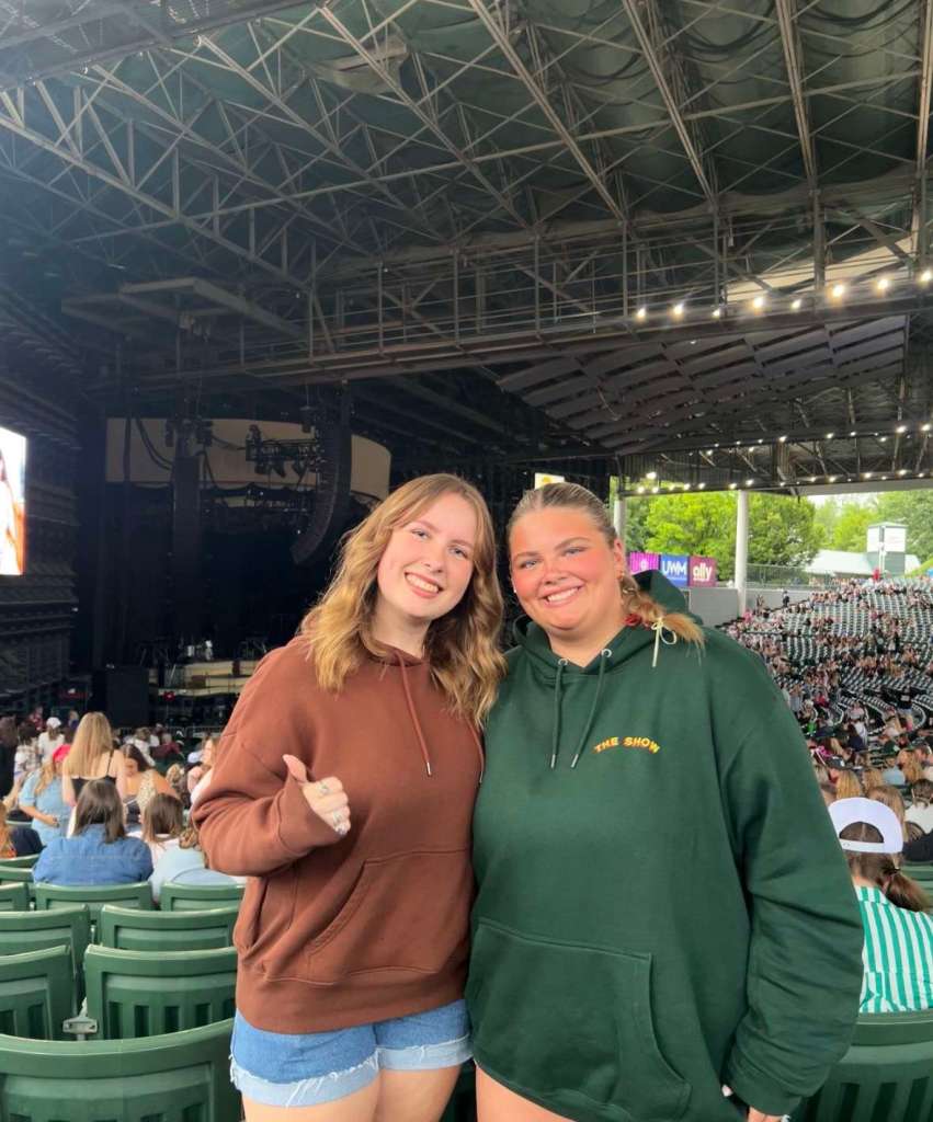 Two women smile and pose together inside an open-air concert venue. One wears a brown hoodie and denim shorts, the other a dark green hoodie with “THE SHOW” written on it. Behind them, rows of green seats are filled with people waiting for the performance to start.