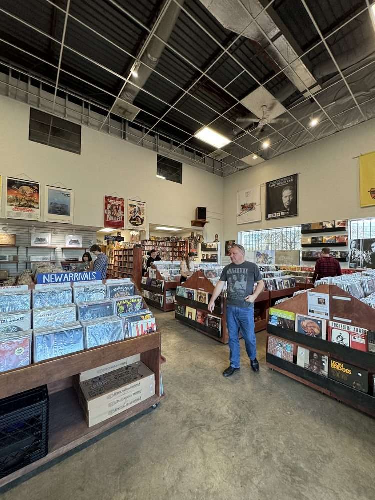 Spacious record store interior with high ceilings, hanging posters, and rows of wooden bins filled with vinyl records. A “New Arrivals” section is displayed in the foreground, while several people browse records throughout the store. The atmosphere feels open and well-organized.