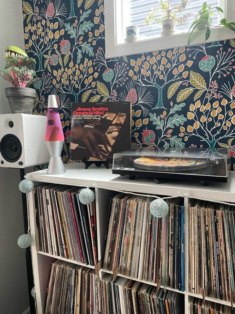 A well-organized vinyl listening corner with a white cube record shelf packed with LPs, a U-Turn turntable, white speakers, a pink lava lamp, and a copy of Jimmy Smith’s Greatest Hits! displayed proudly. The space is framed by bold, botanical wallpaper and natural light from a small window above.