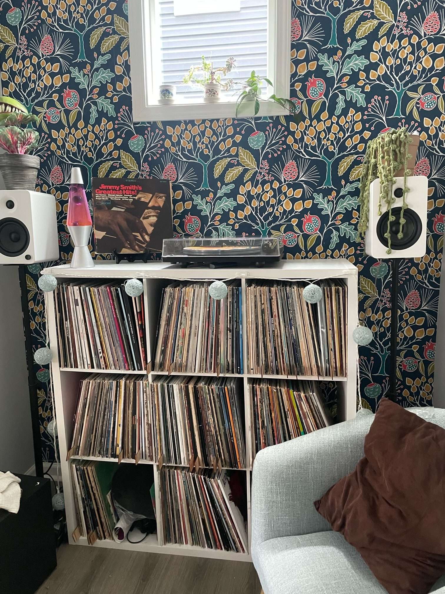 Full setup view of a cozy vinyl nook featuring a turntable, speakers, lava lamp, and a lush mix of potted plants. The colorful floral wallpaper, teal armchair, and record-filled shelf create a vibrant and inviting listening space.