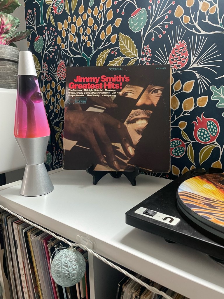 Tight close-up of Jimmy Smith’s Greatest Hits! album on a black stand next to a lava lamp and turntable. The colorful wallpaper in the background adds character to the shelf filled with vinyl records below.