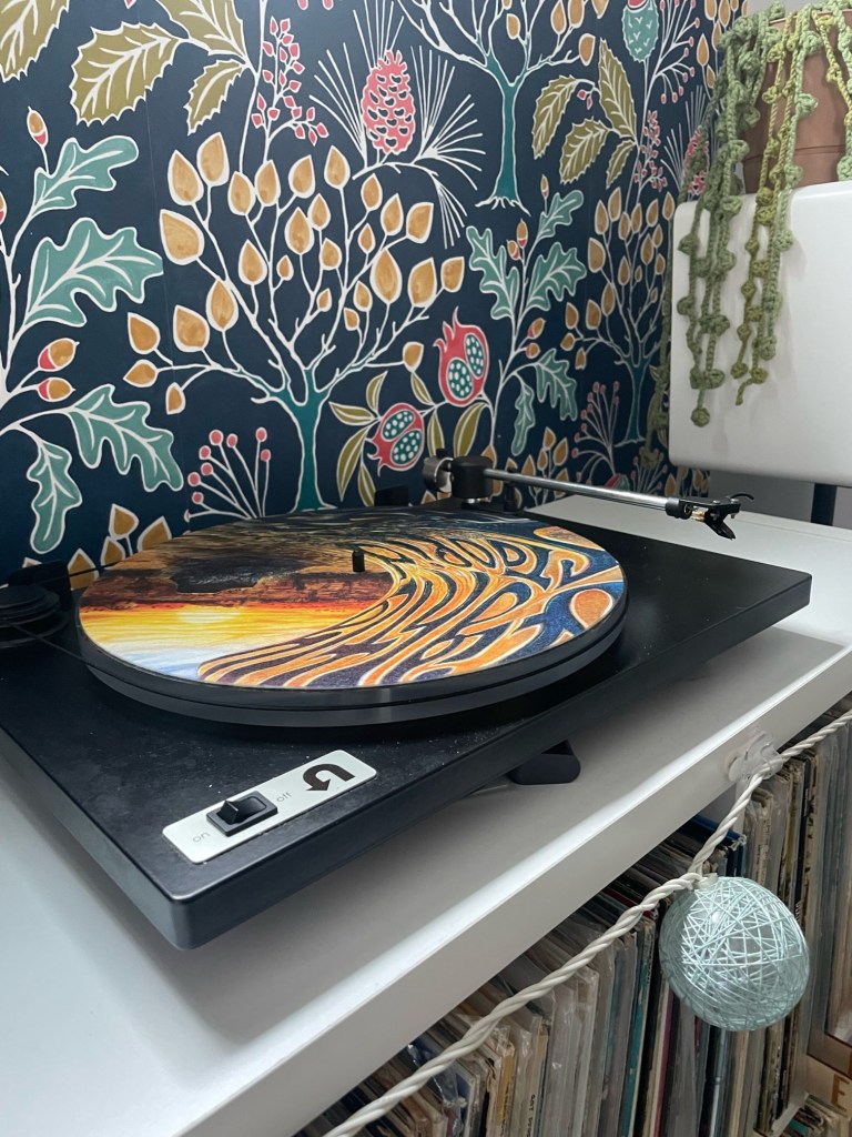 Side angle of a U-Turn turntable featuring a vibrant, psychedelic slipmat. The tonearm is positioned above the slipmat, with a speaker and cascading potted plant in the background. String lights and vinyl records sit neatly below the setup.