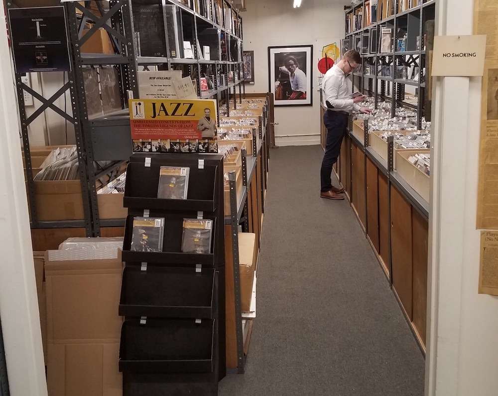 A man in a white shirt browses through tightly packed rows of jazz records inside a narrow shop lined with metal shelving. Boxes and vinyl sleeves fill the shelves, and a display stand at the entrance showcases jazz CDs. A framed black-and-white photo of a musician hangs on the back wall, adding to the archival, collector’s vibe of the space.