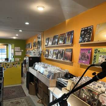 Interior of Love Wheel Records, a small record shop with orange and green painted walls. Vinyl records are displayed on wall-mounted shelves and in bins below. A rug covers part of the floor, and a microphone stand is visible in the foreground, hinting at possible in-store performances.