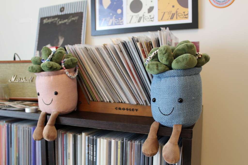 Two smiling plush plant pots sit on top of a Crosley record shelf filled with vinyl, with a Marshall speaker and framed artwork in the background.