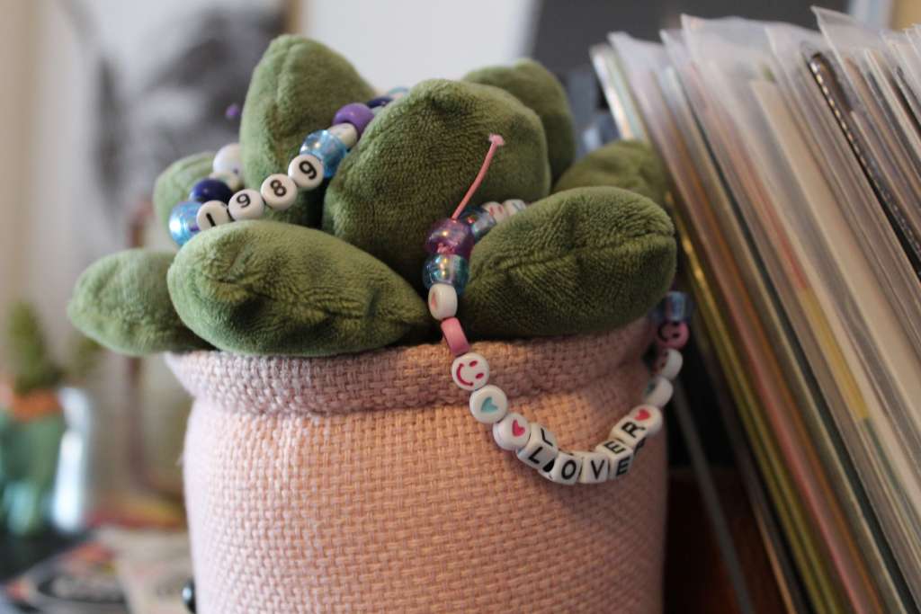Detail shot of a pink plush plant pot with a handmade bead bracelet reading “1989” and “Lover” draped across the leaves.