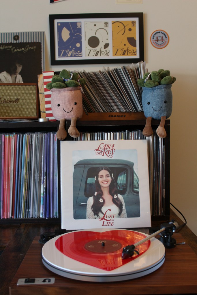Vinyl record setup with Lana Del Rey’s “Lust for Life” album displayed on a shelf behind a wooden turntable spinning a red-and-white vinyl record. Surrounding the setup are shelves of records, a Marshall speaker, and two smiling plant plushies.