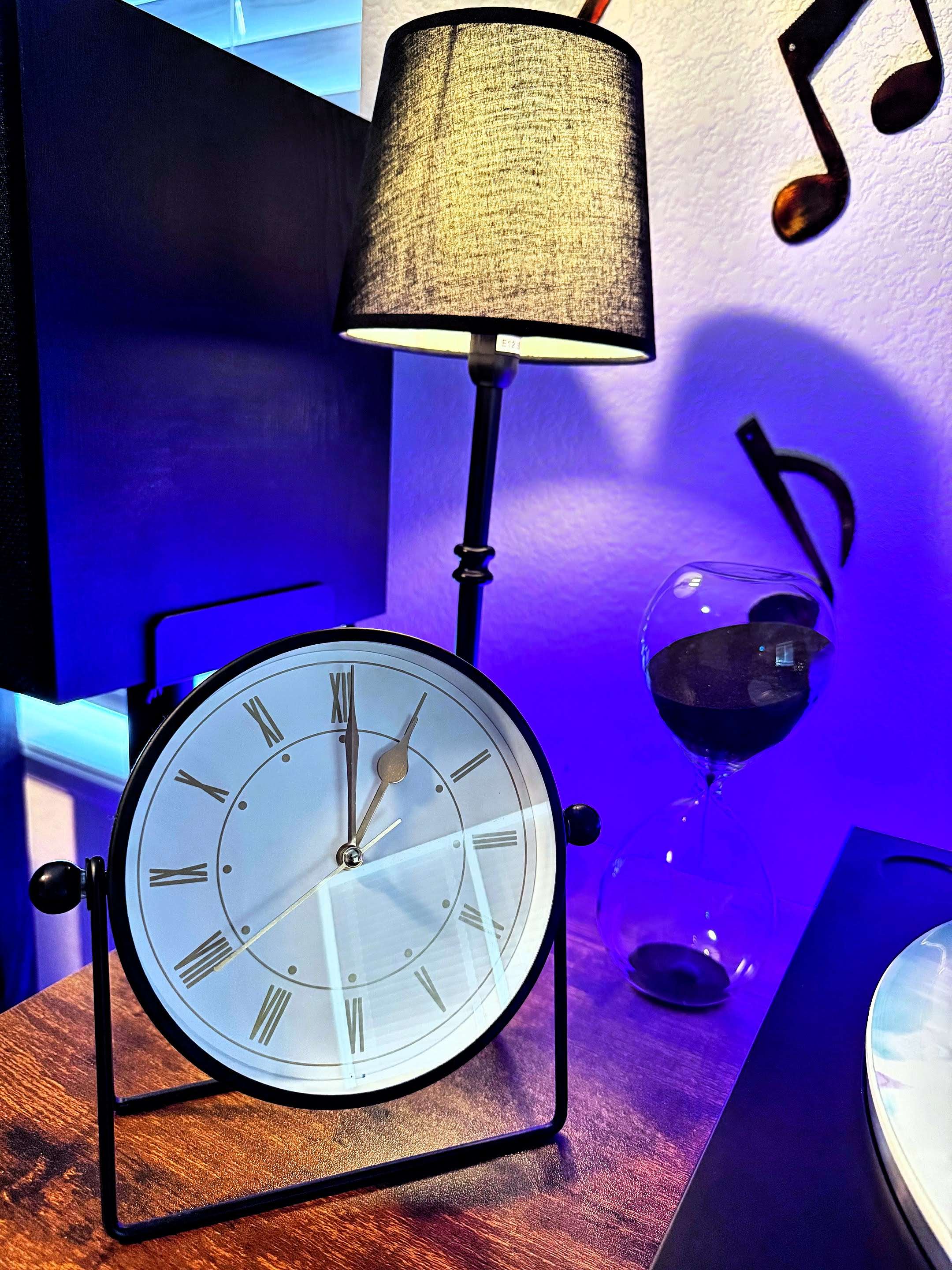 A tabletop setup with a black-framed clock showing Roman numerals, a black lamp with a fabric shade, and a glass hourglass filled with black sand. A wall decorated with music note cutouts and blue accent lighting creates a cozy atmosphere.