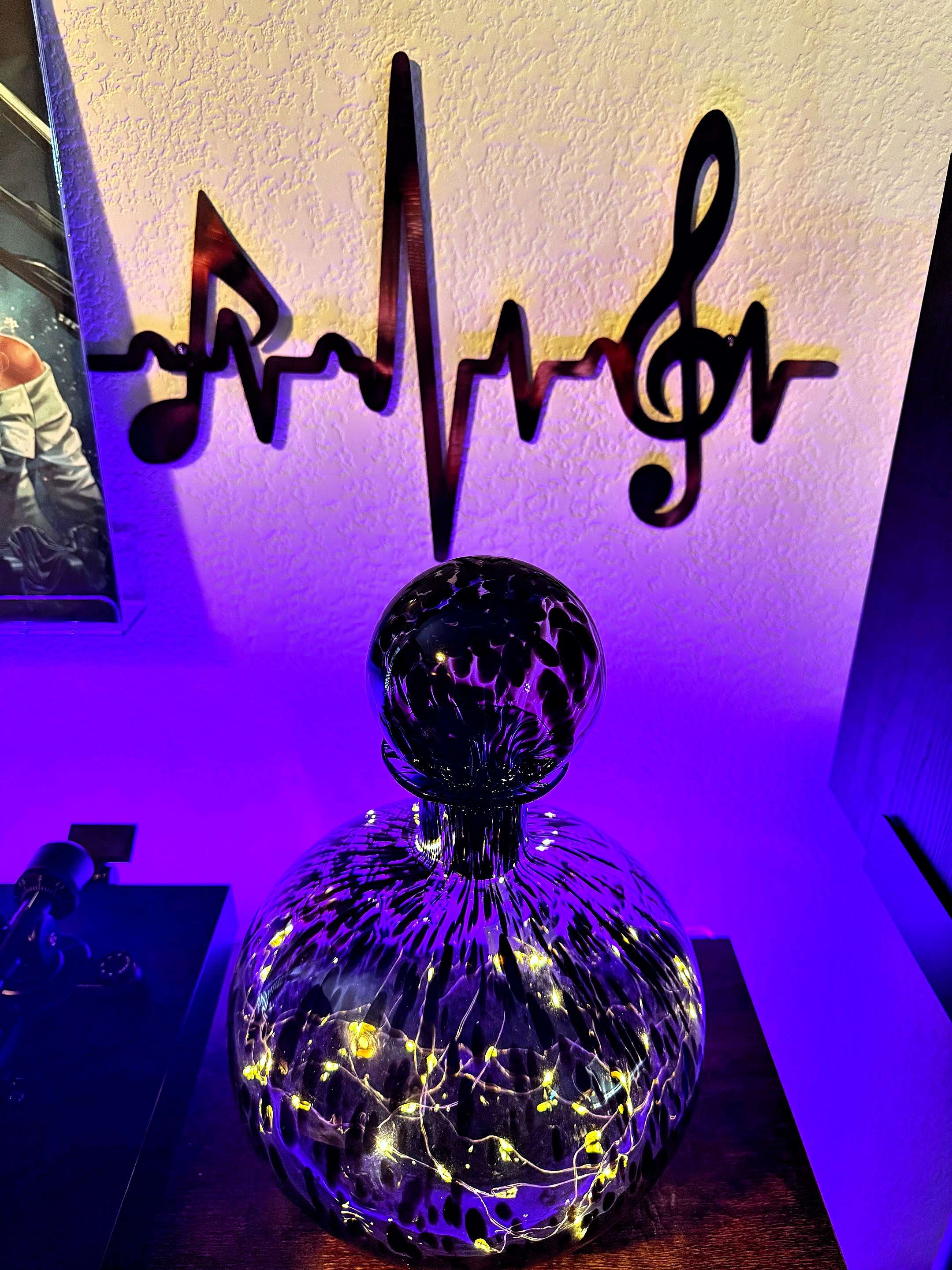 A decorative glass lamp filled with string lights glows warmly against a wall. Above it, a metal wall art piece shaped like a musical heartbeat with notes and a treble clef is mounted. Purple-blue accent lighting enhances the scene.
