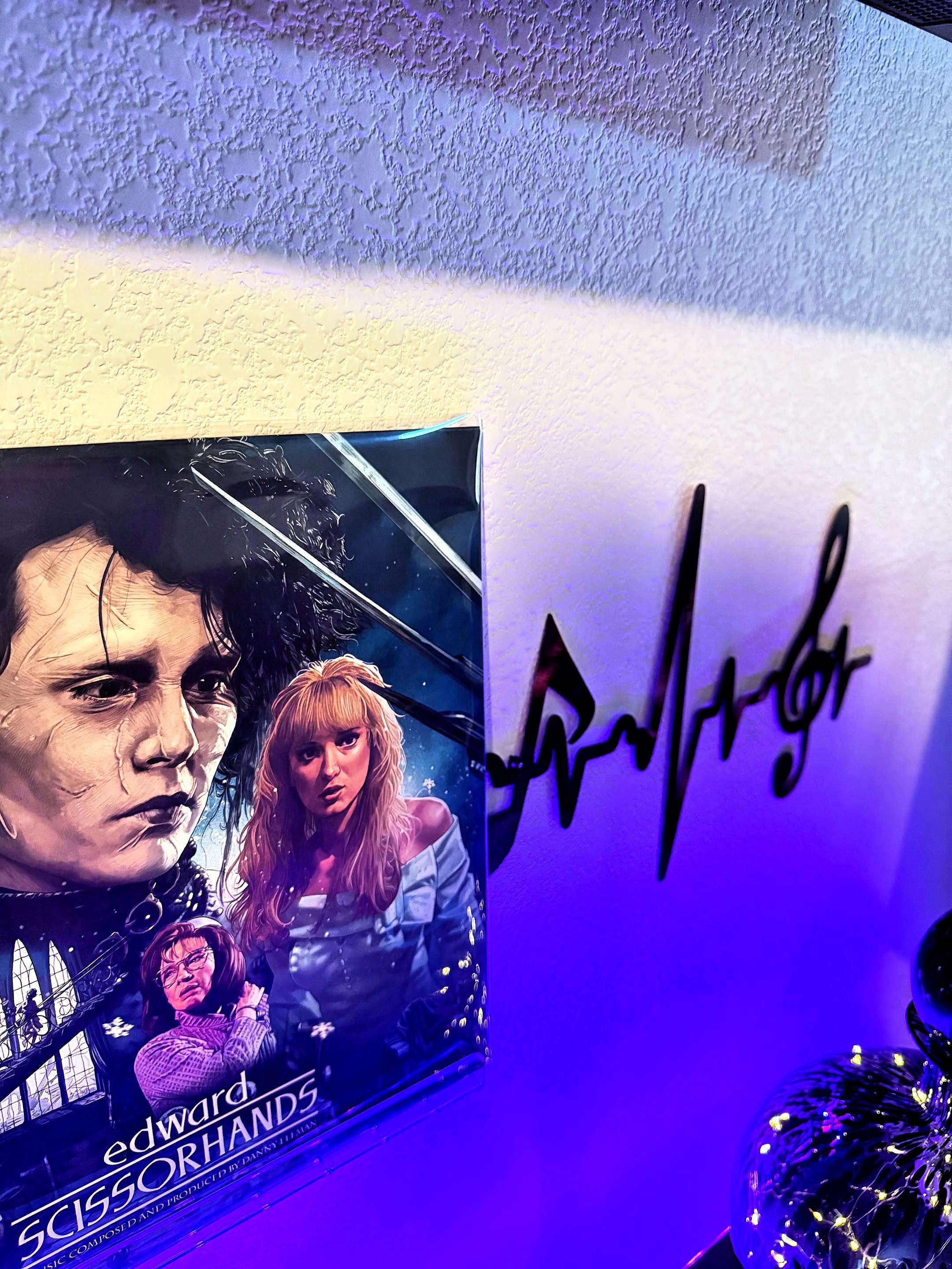 A framed Edward Scissorhands movie poster displayed on a wall, with part of a treble clef heartbeat wall art design visible beside it. Blue-purple lighting casts a dramatic glow across the textured wall.