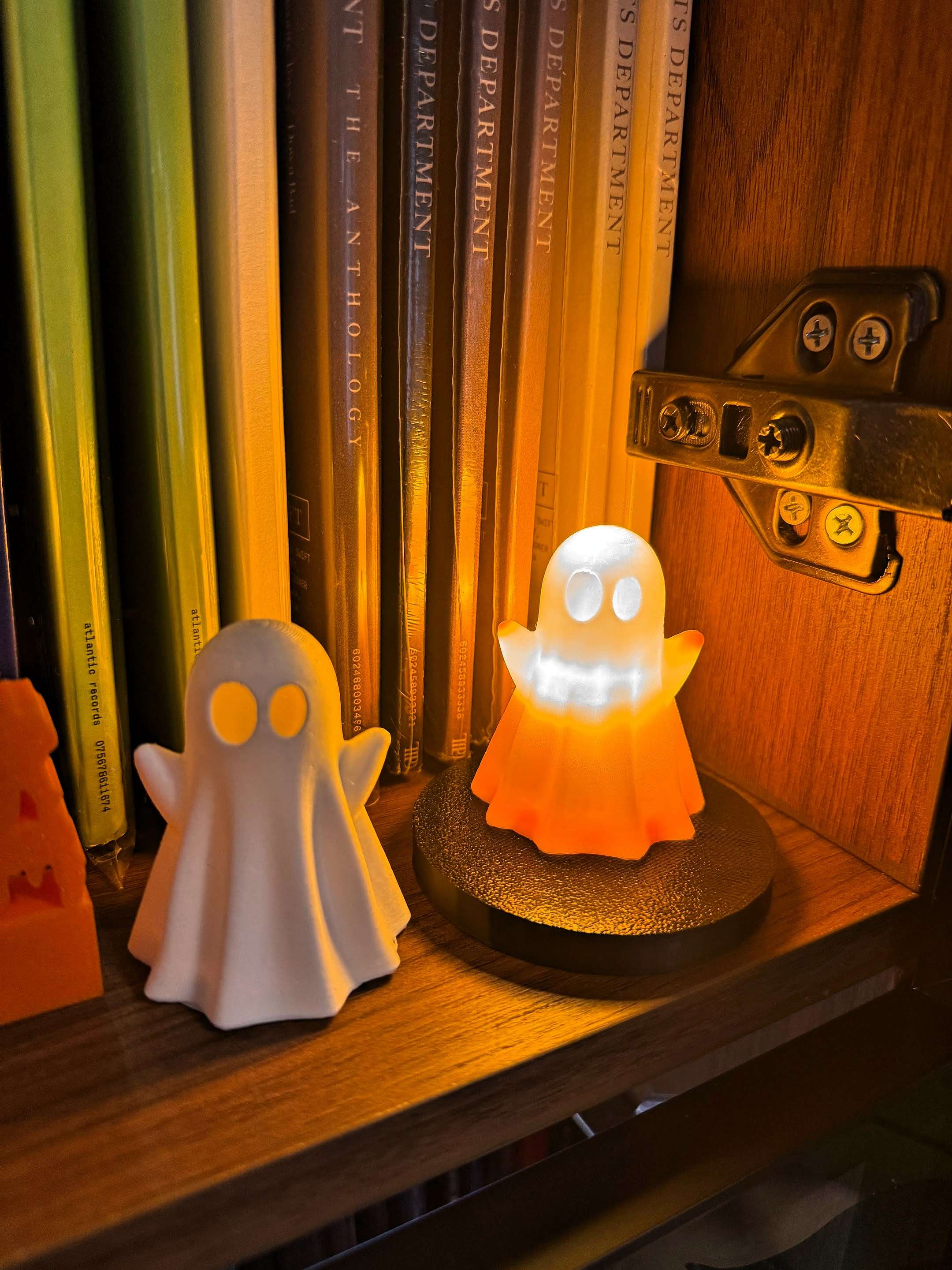 Two small ghost figurines on a wooden bookshelf, one white and one orange that lights up. They sit in front of neatly arranged records and books, adding a playful Halloween touch.