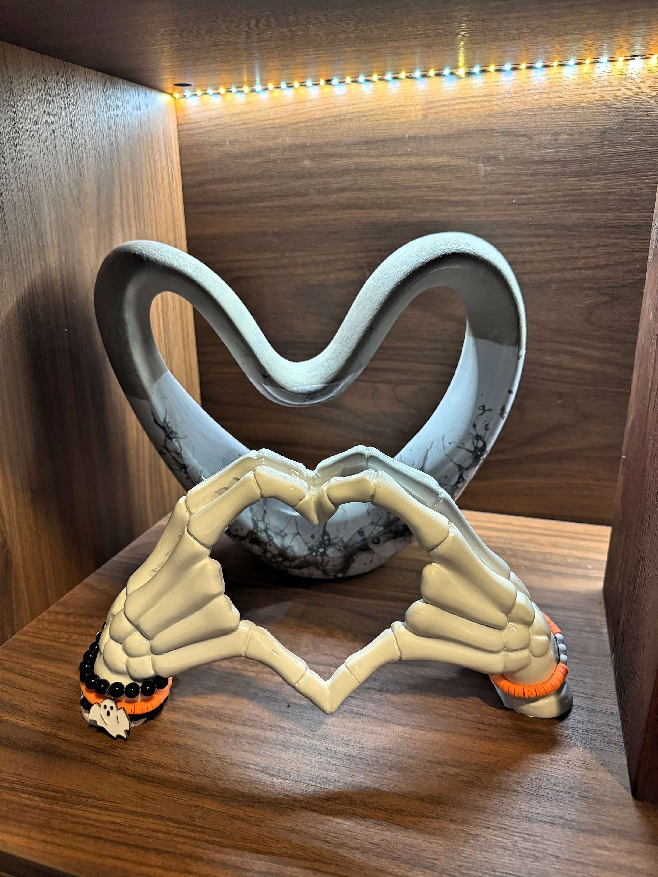 A Halloween decoration featuring two skeleton hands forming a heart shape, set on a wooden shelf. Behind them is a gray, abstract heart-shaped ornament, with warm LED strip lights above.