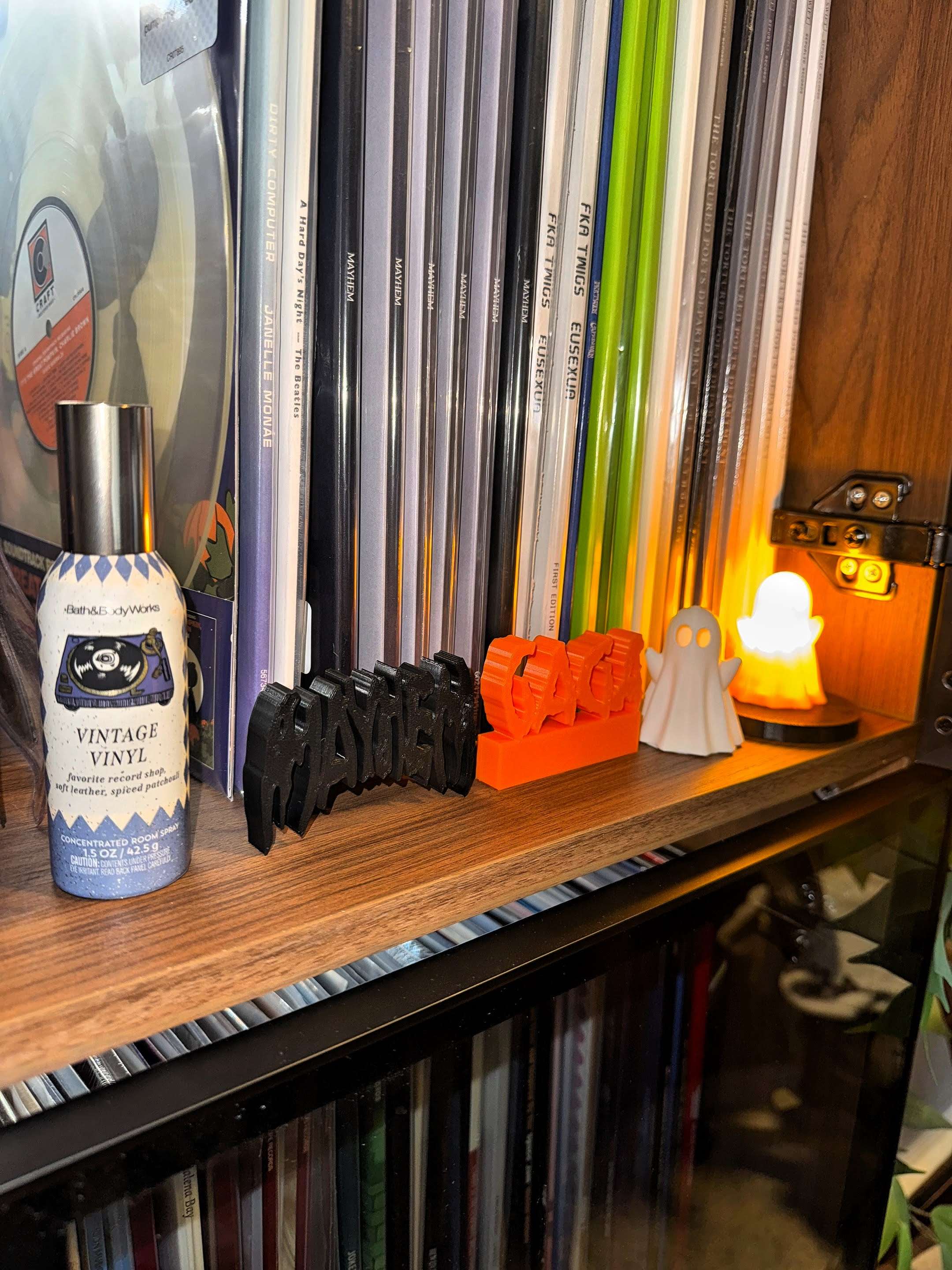 A shelf lined with vinyl records, decorated with small Halloween-themed ornaments including a ghost, jack-o’-lanterns, and a black “HAUNTED” sign. A bottle labeled “Vintage Vinyl” spray sits beside the decorations, lit by a small glowing orange ghost lamp.