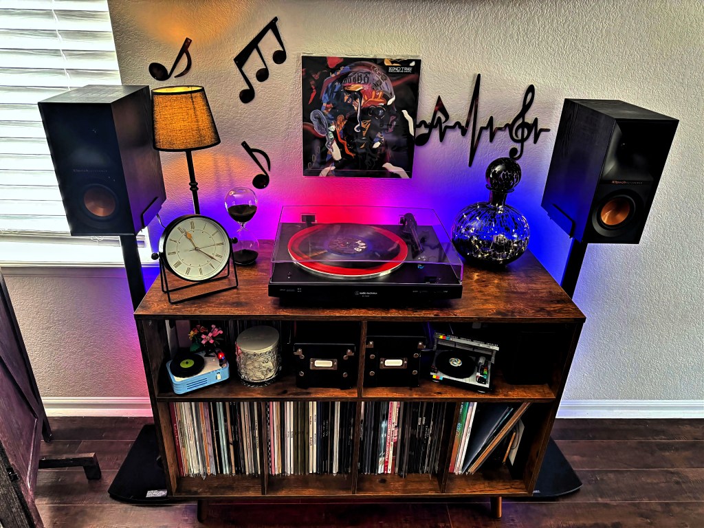 Wide shot of a modern vinyl listening station with wooden console, Klipsch speakers on stands, LED backlighting, and artistic wall decor including music notes and a heartbeat treble clef. The setup includes a turntable, wine hourglass, decorative decanter, and an analog table clock.