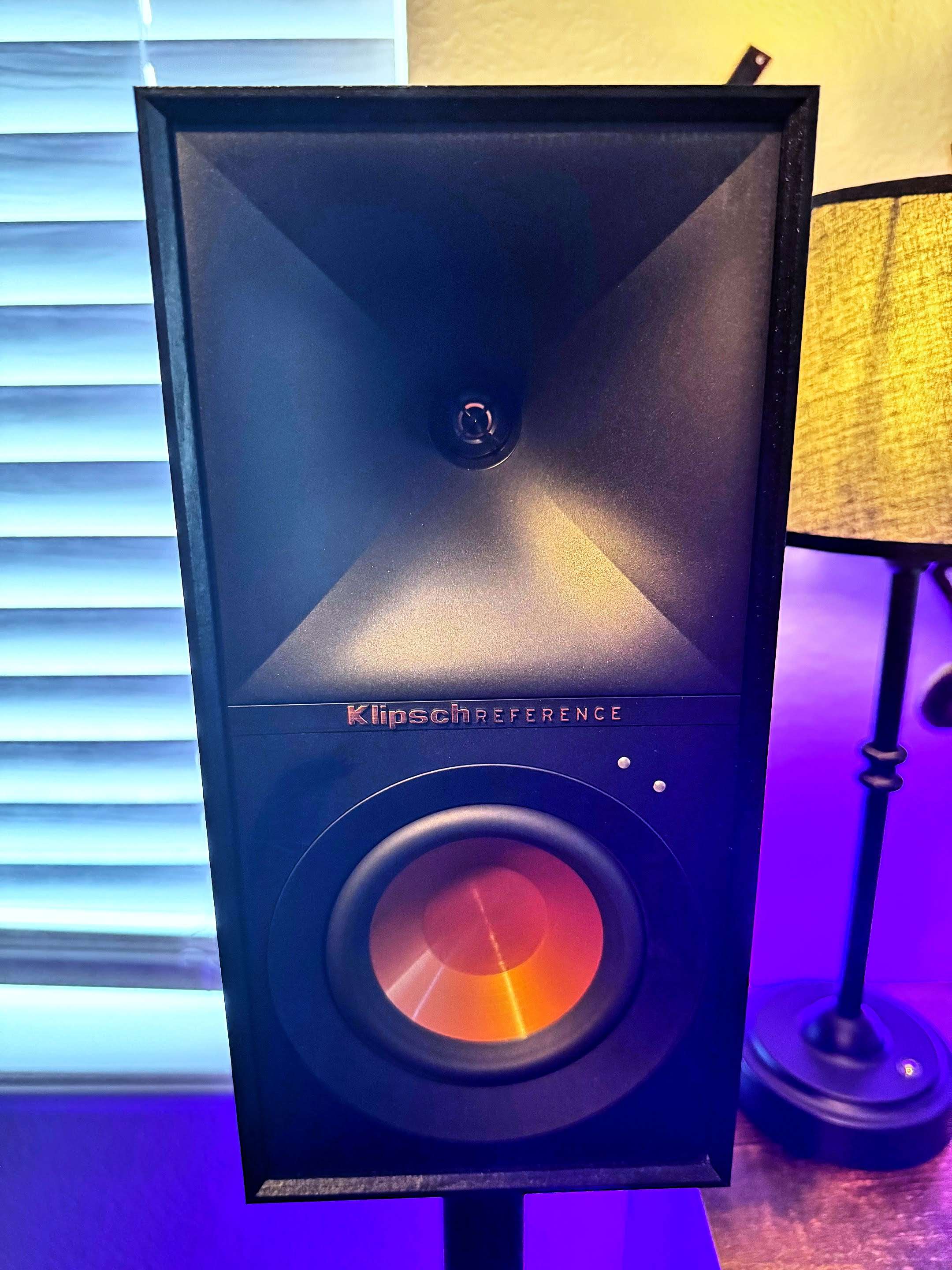 Close-up of a Klipsch Reference speaker with its signature copper-colored woofer and black horn tweeter design. The speaker sits on a stand next to a lamp, with blue and yellow lighting adding a warm glow to the setup.