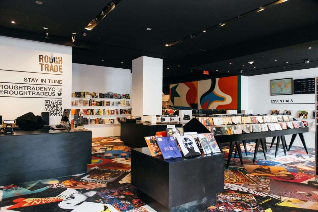 Interior of Rough Trade record store in New York City, featuring sleek black display tables filled with vinyl records, colorful wall art in the background, and a patterned floor covered in album artwork. The checkout counter is on the left, with books and music merch displayed along the walls.
