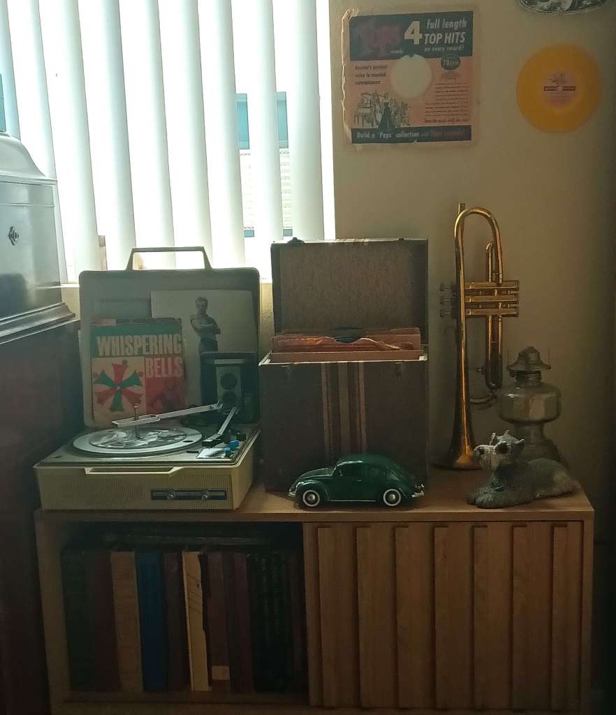 Vintage suitcase-style record player with a box of 45s, trumpet, lamp, and decor on a mid-century cabinet.