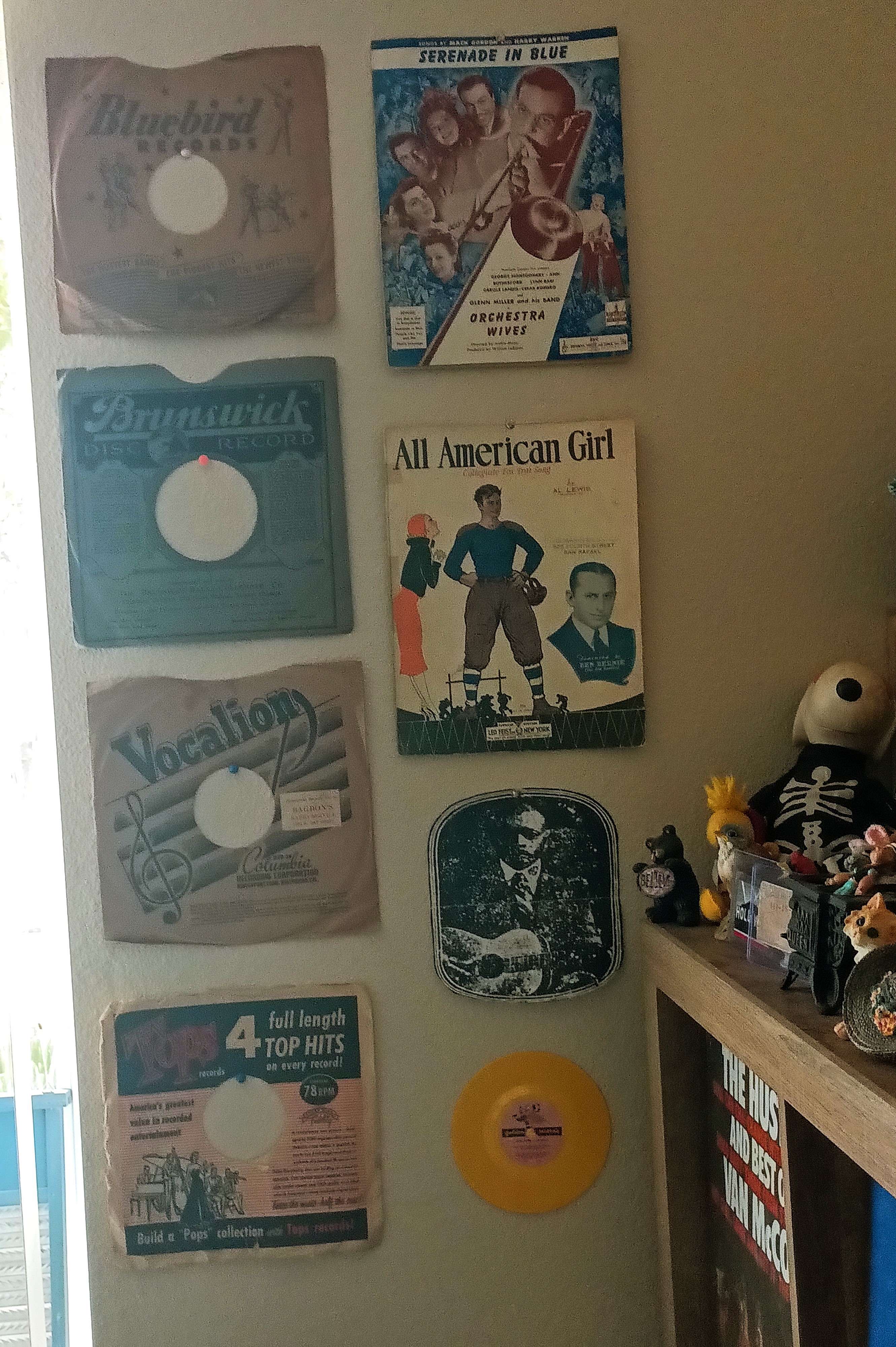 Gallery wall with vintage 78/45 record sleeves, sheet music, and small collectibles near a record shelf.