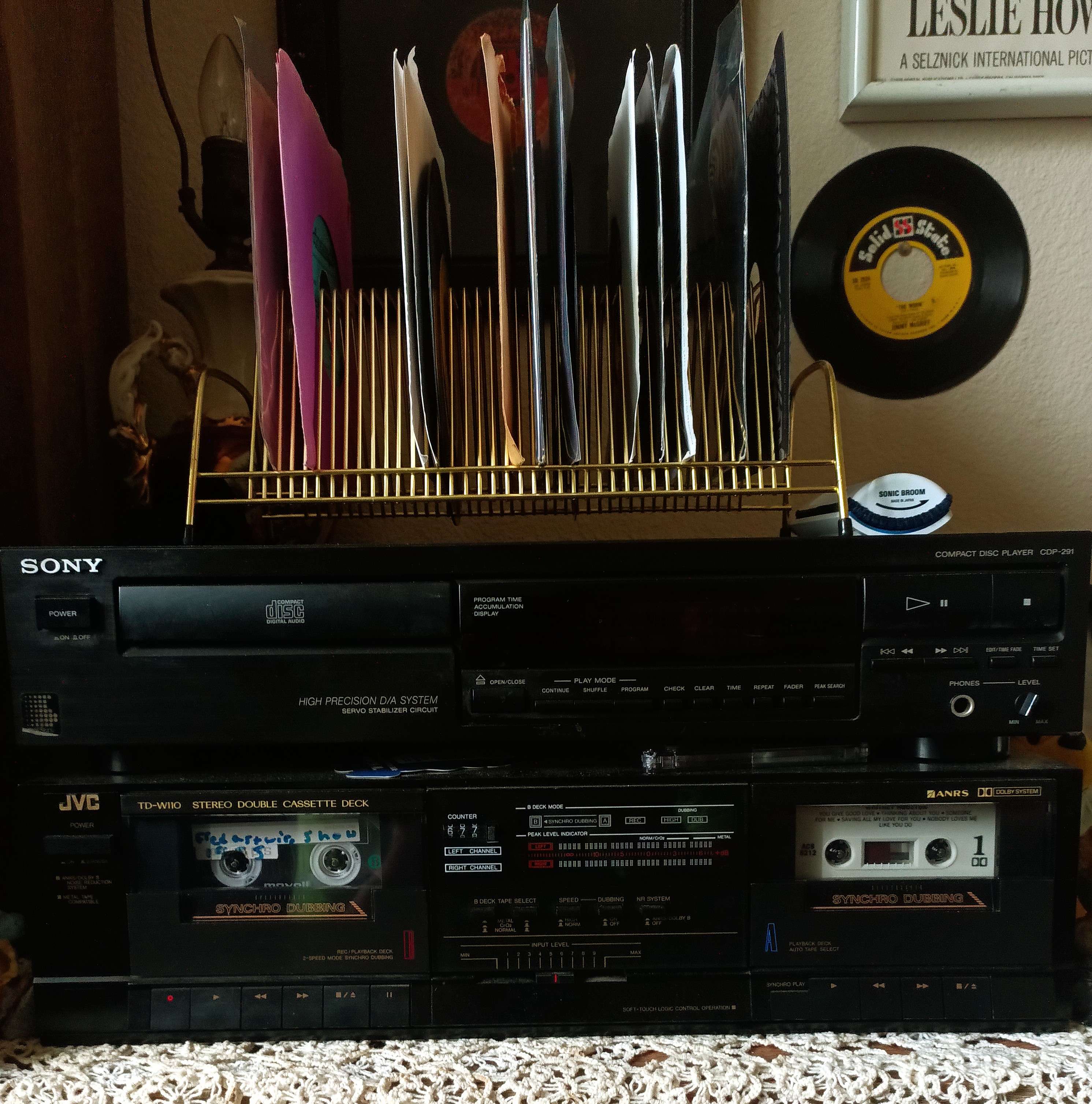 Sony CD player and JVC double cassette deck stacked, with a gold rack holding colorful 45-rpm records above.