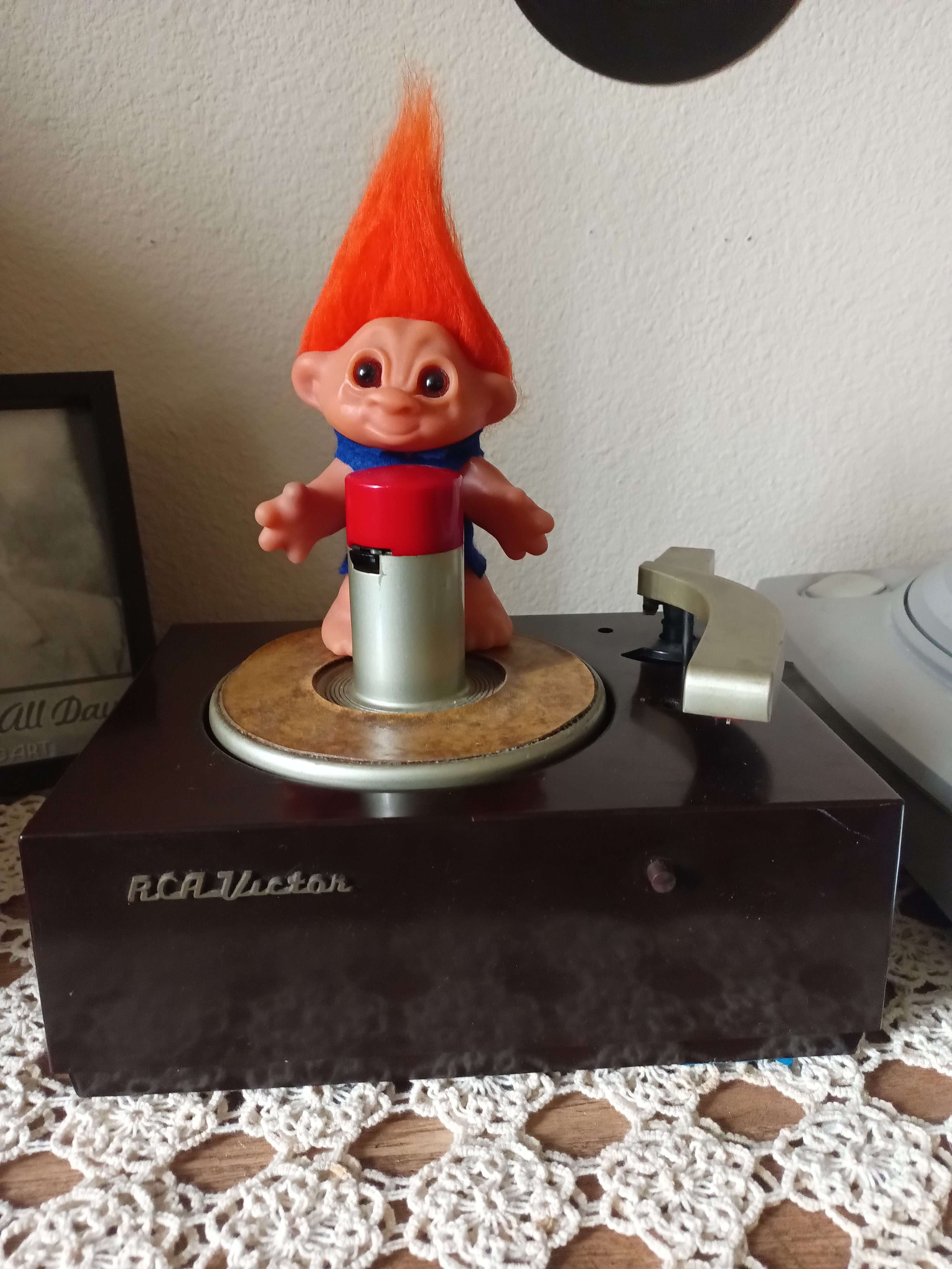 Wallace the vintage troll mascot perched on an RCA Victor 45-rpm record player on a lace-covered table.
