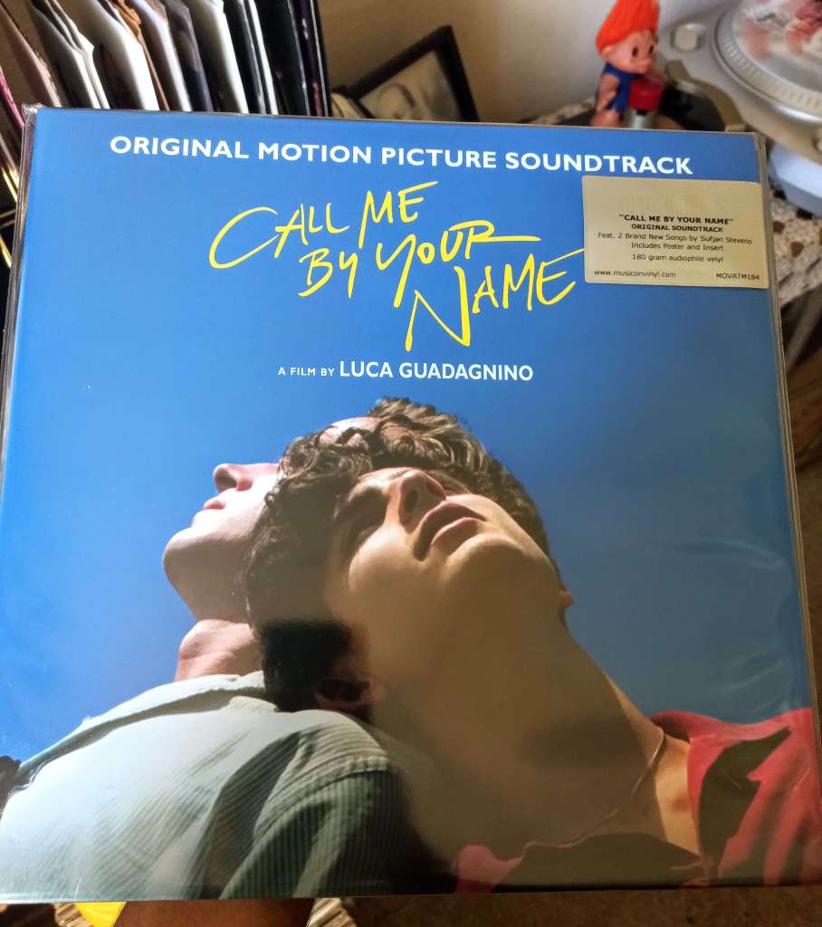 The blue-toned album cover for Call Me By Your Name, showing two men leaning their heads back under bright sunlight with yellow handwritten text for the title.