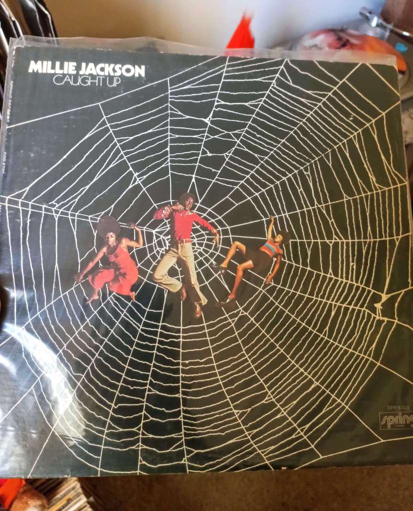 The album cover for Caught Up by Millie Jackson, showing three people entangled in a giant white spiderweb against a black background, symbolizing being “caught up.”