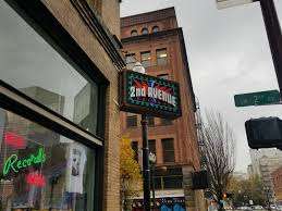 Street view of 2nd Avenue Records in downtown Portland, Oregon, with its neon sign hanging above the sidewalk. The record shop sits in a brick building surrounded by classic Portland architecture on a cloudy day.