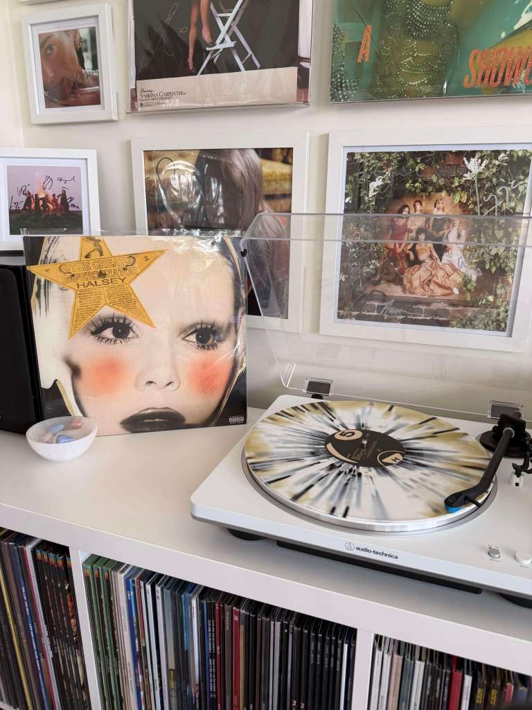 The Great Impersonator” on cream/black splatter vinyl spinning on a white Audio-Technica turntable, framed wall prints behind.