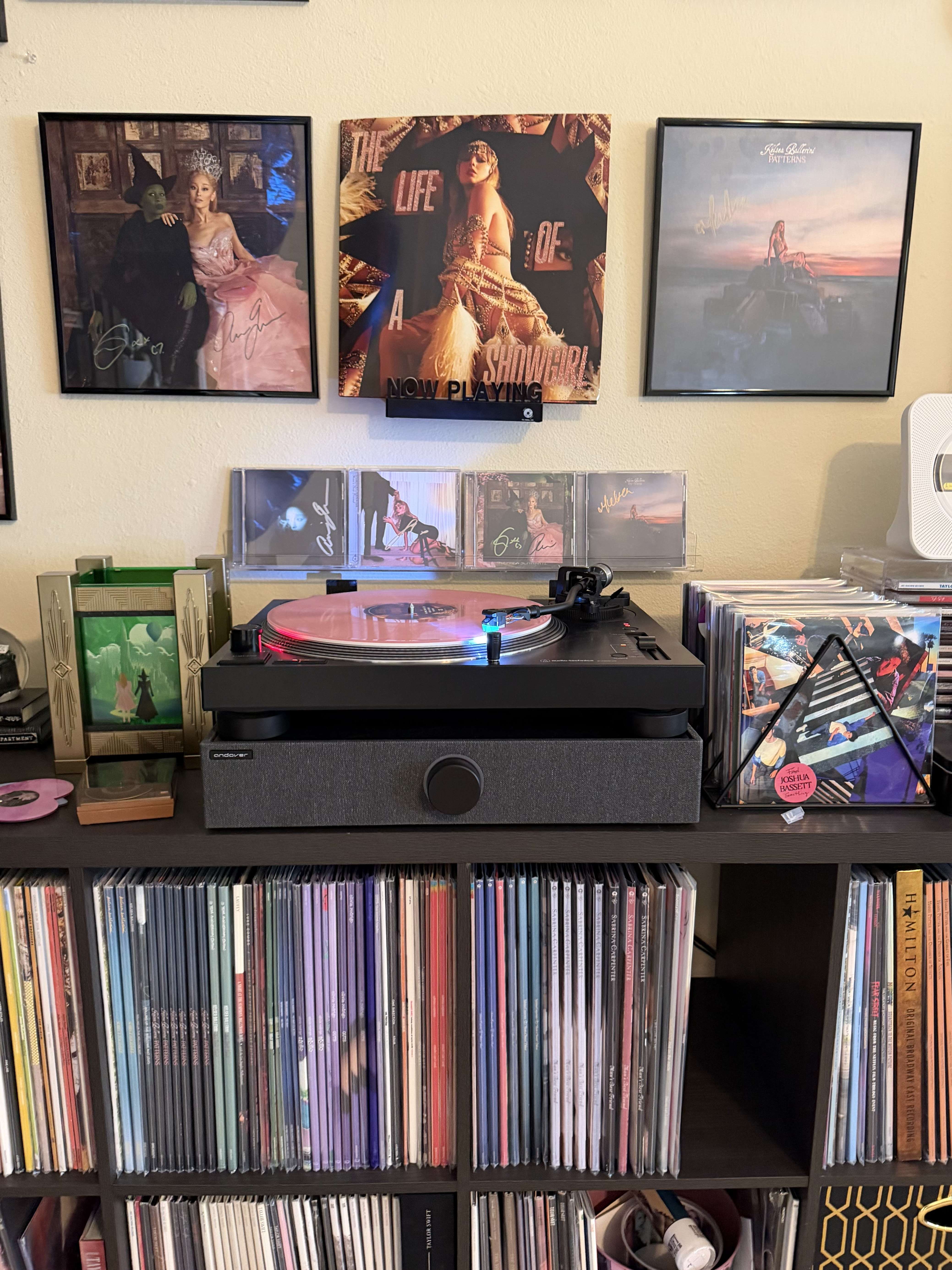 Close-up of the turntable on a black console with a pink vinyl spinning, LP rows in cubbies, and small display items nearby.