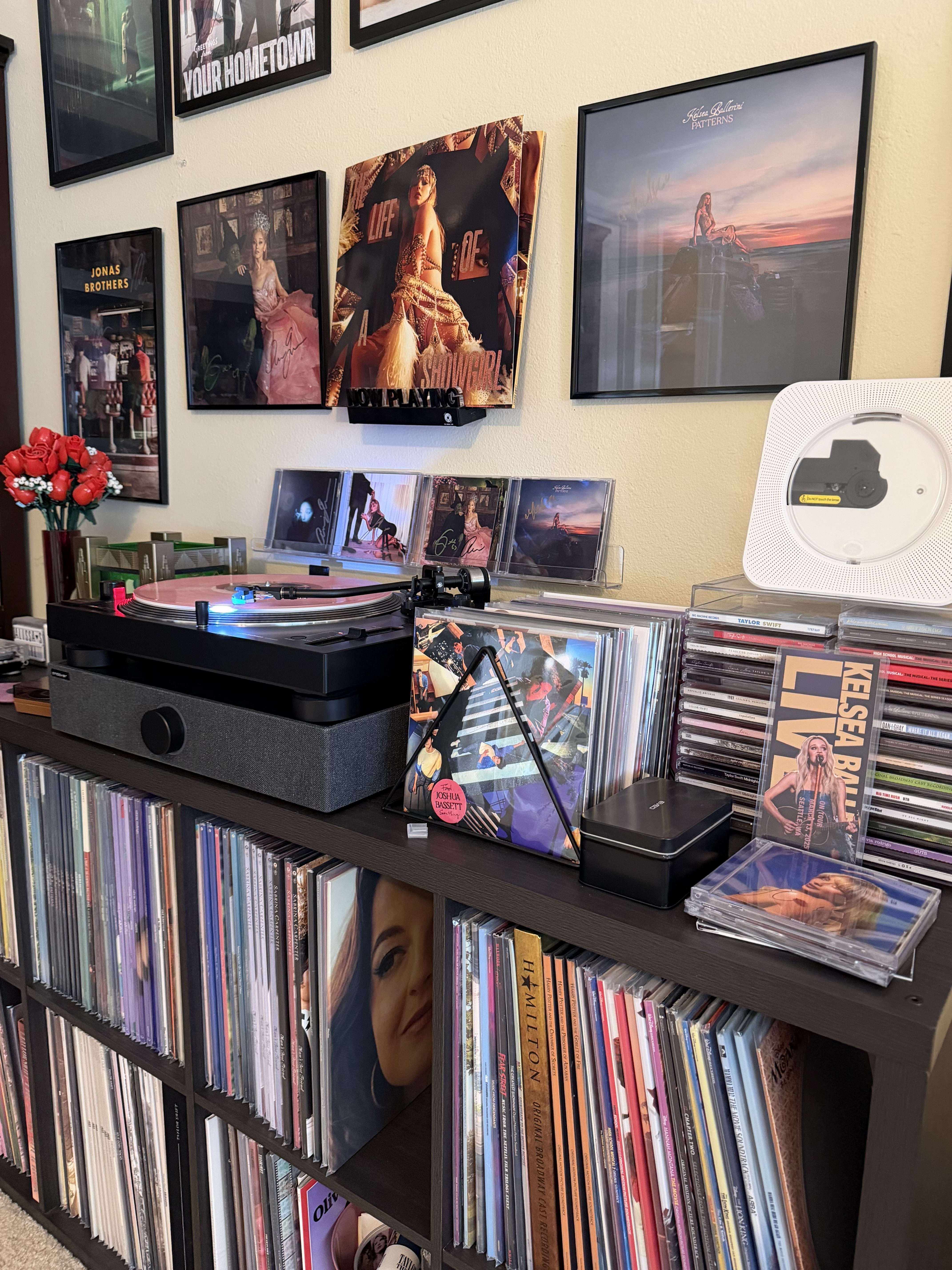 Angled close-up of the console: turntable spinning a pink record, signed CDs, a triangle now-playing stand, and rows of LPs below.