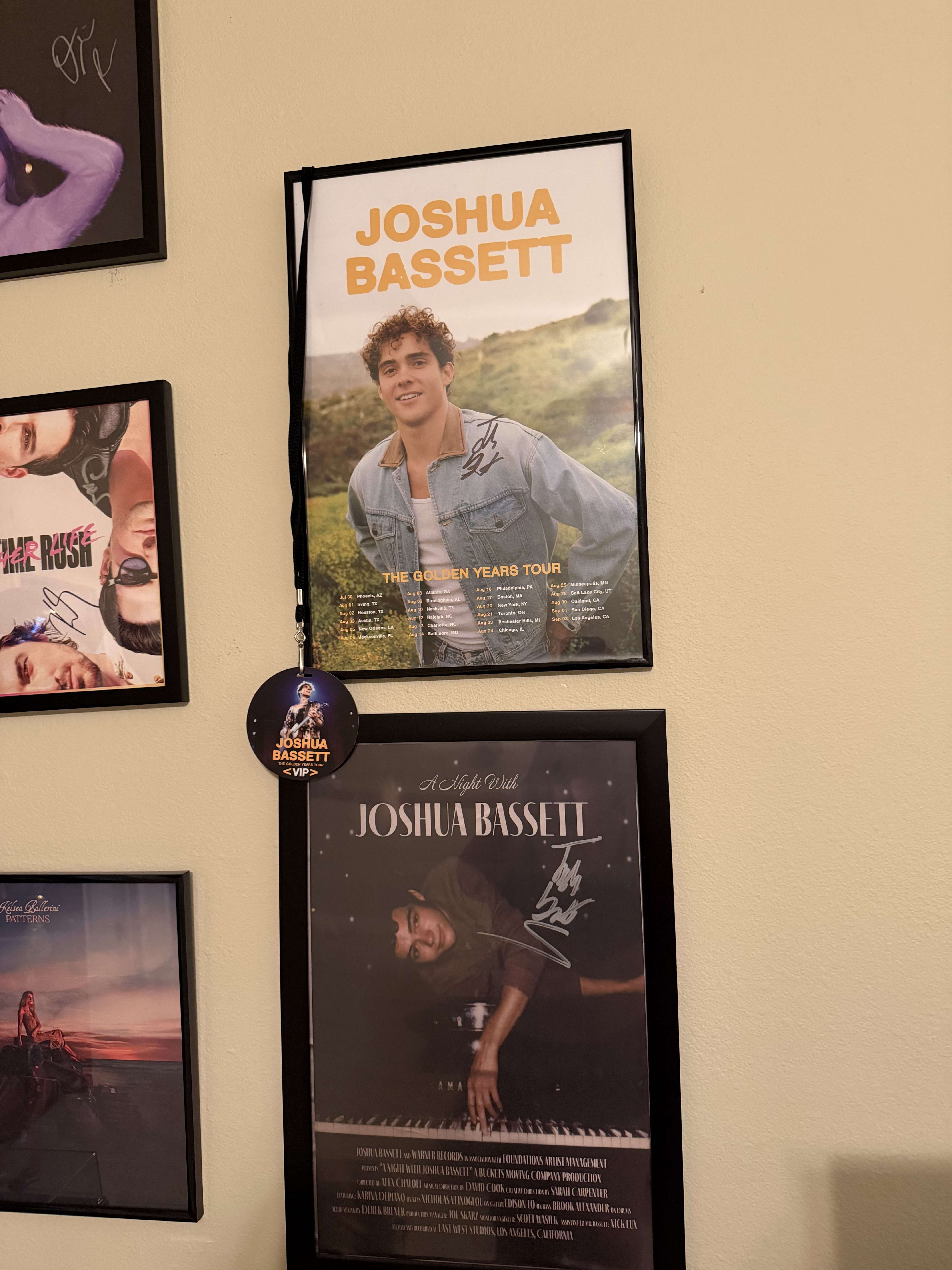 Two framed Joshua Bassett posters hanging on a beige wall — one for The Golden Years Tour and another for A Night With Joshua Bassett — both autographed, with a VIP badge hanging between them.