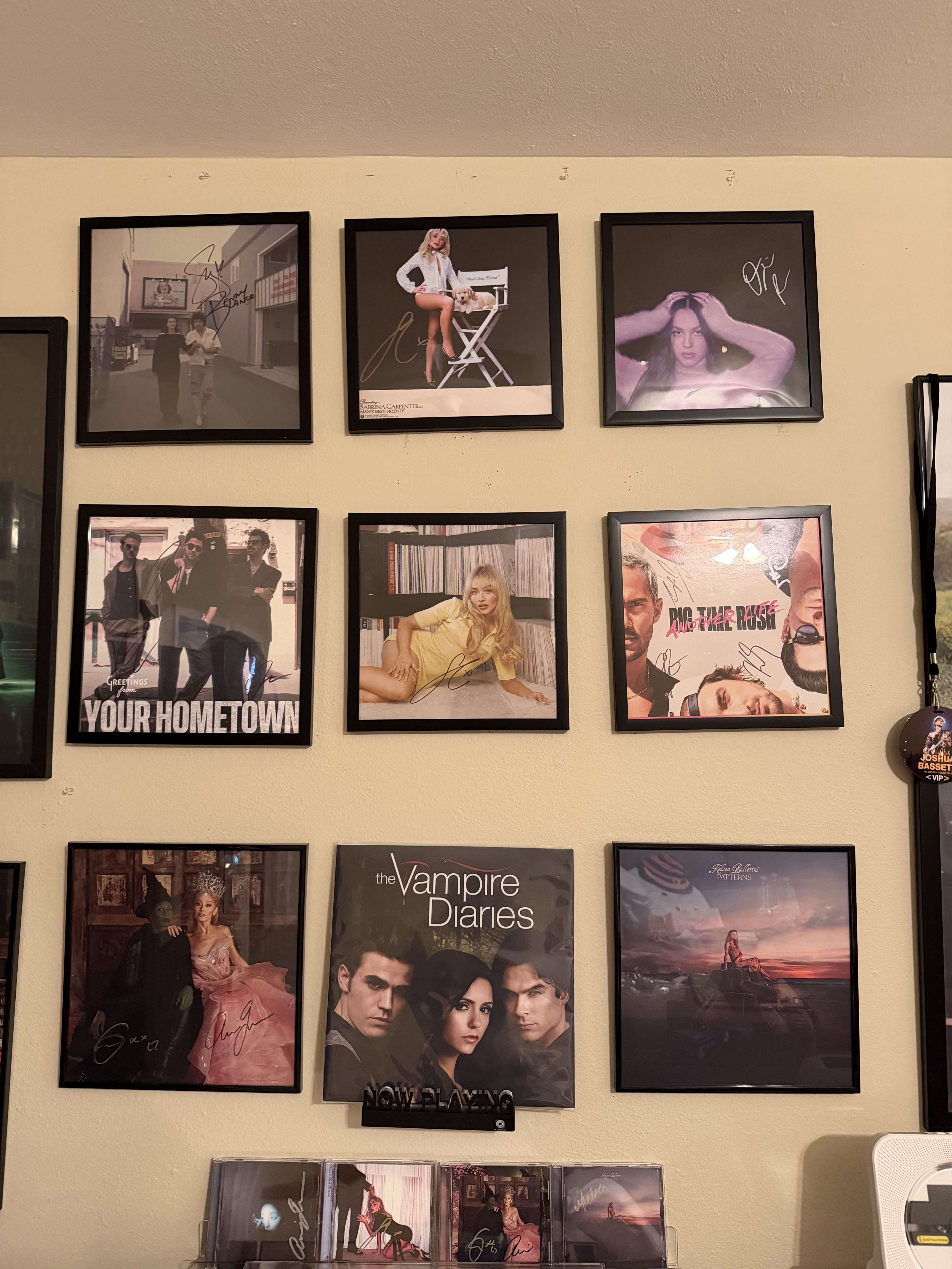 A grid of framed album covers and posters arranged on a wall, featuring autographed vinyls from various artists and shows, including The Vampire Diaries, Wicked, and Your Hometown.