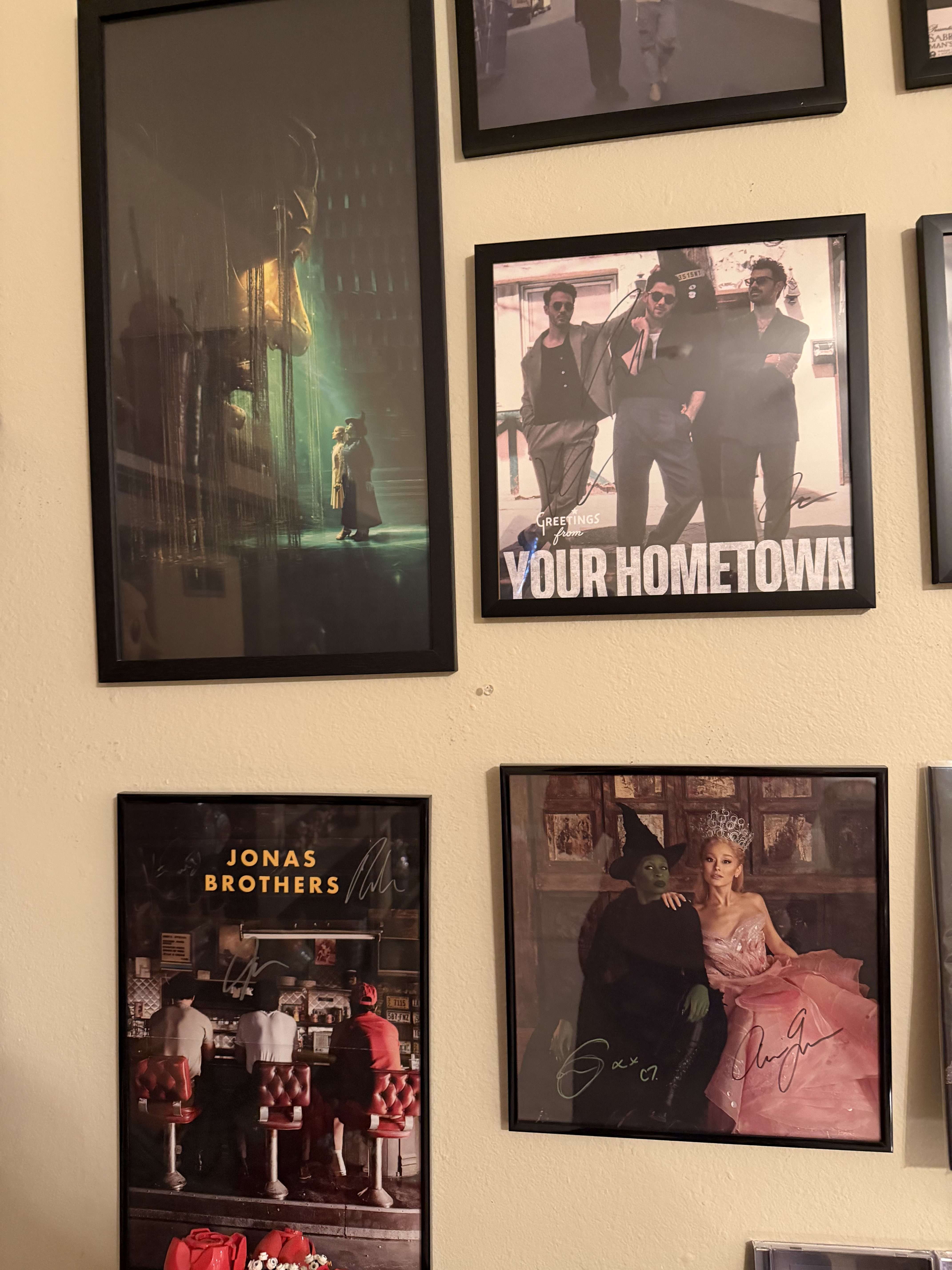 A closer shot of framed posters on a wall, including Jonas Brothers, Your Hometown, and Wicked, surrounded by other music and pop culture art pieces.
