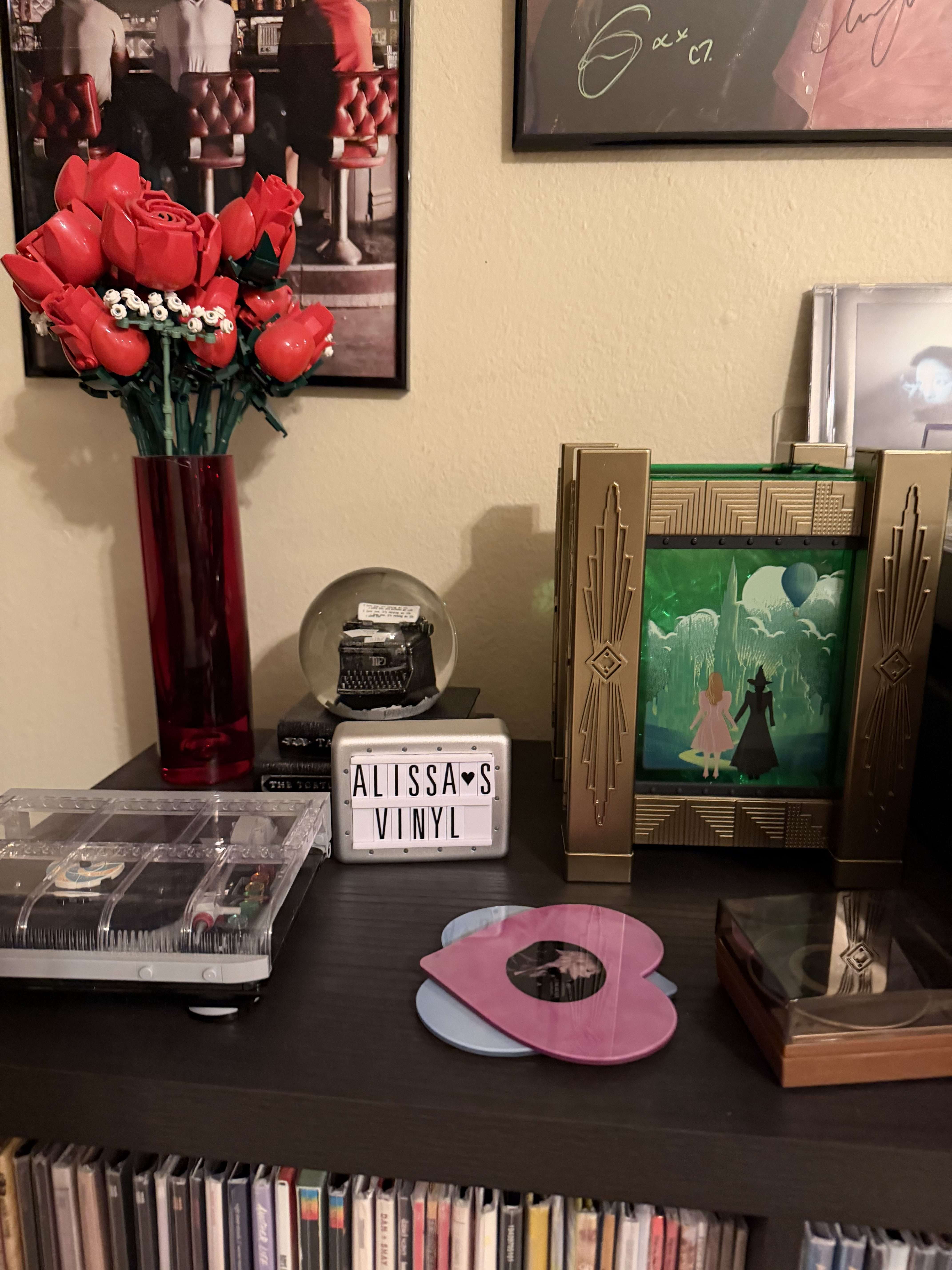 A cozy display shelf featuring a LEGO record player, a vase with red roses, a framed Wicked artwork, a sign reading “Alissa’s Vinyl,” and colorful heart-shaped vinyl records laid out on the surface.