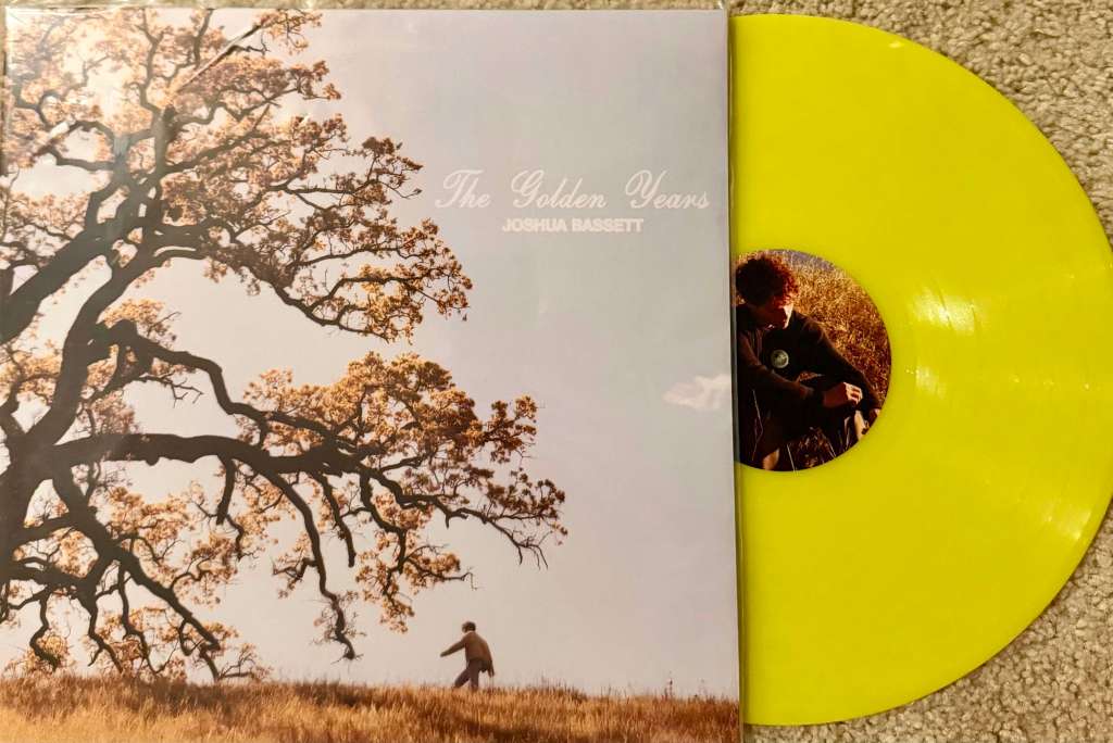 Joshua Bassett’s “The Golden Years” vinyl record displayed with its yellow pressing partially pulled from the sleeve, which features a scenic photo of a tree and a man walking through a golden field.