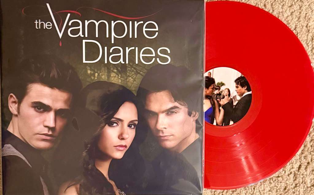 The Vampire Diaries soundtrack vinyl featuring the three main characters on the cover and a translucent red record partially pulled out, with a label image of a dance scene from the show.
