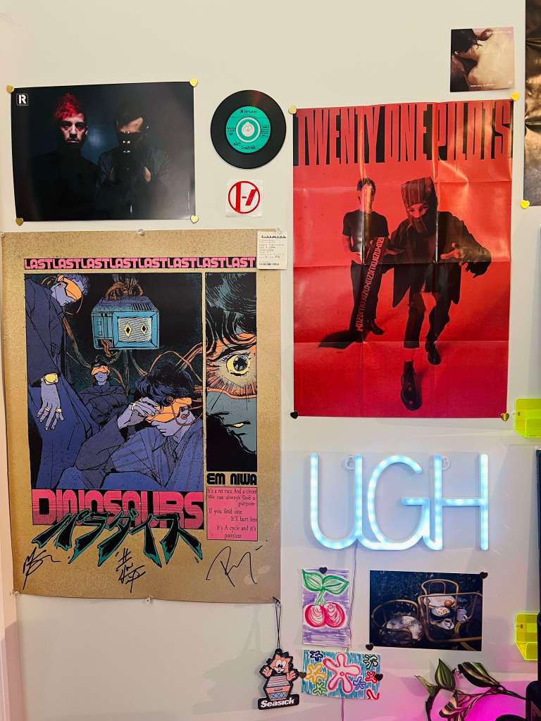 Wall collage with Twenty One Pilots posters, a small 7-inch record, a signed ‘Dinosaurs’ art print, and a glowing ‘UGH’ neon sign surrounded by mini art cards and trinkets.