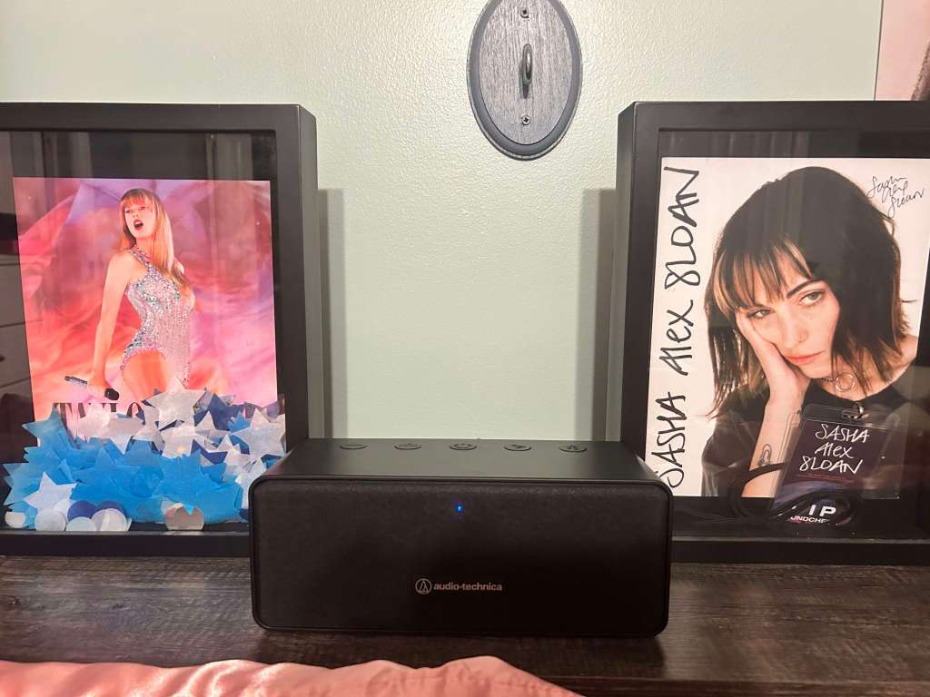 Audio-Technica Bluetooth speaker placed between two framed artist photos on a desk, one showing a stage performance and the other an autographed portrait.
