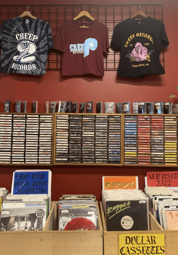 Interior of Creep Records in Philadelphia showing walls lined with cassette tapes, racks of vinyl organized by genre, and T-shirts hanging above labeled “Creep Records.”