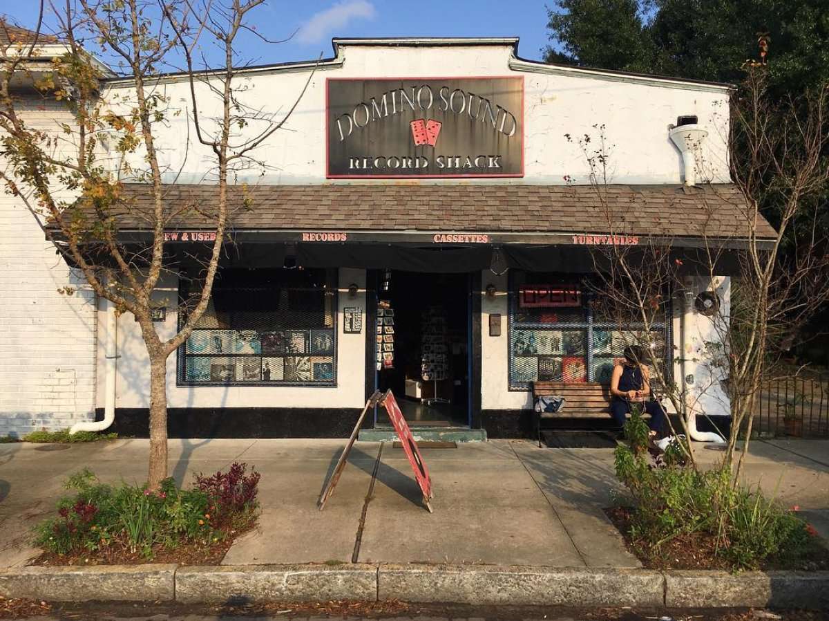 Front view of Domino Sound Record Shack, a cozy neighborhood record store in New Orleans known for reggae, world, and jazz vinyl, with a bench outside and a red sandwich board sign.