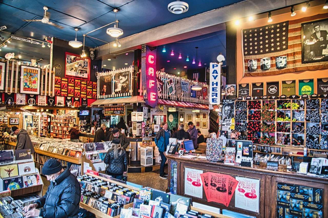 Busy, colorful record shop with a neon “CAFE” sign, posters and merch walls, and customers flipping through bins.