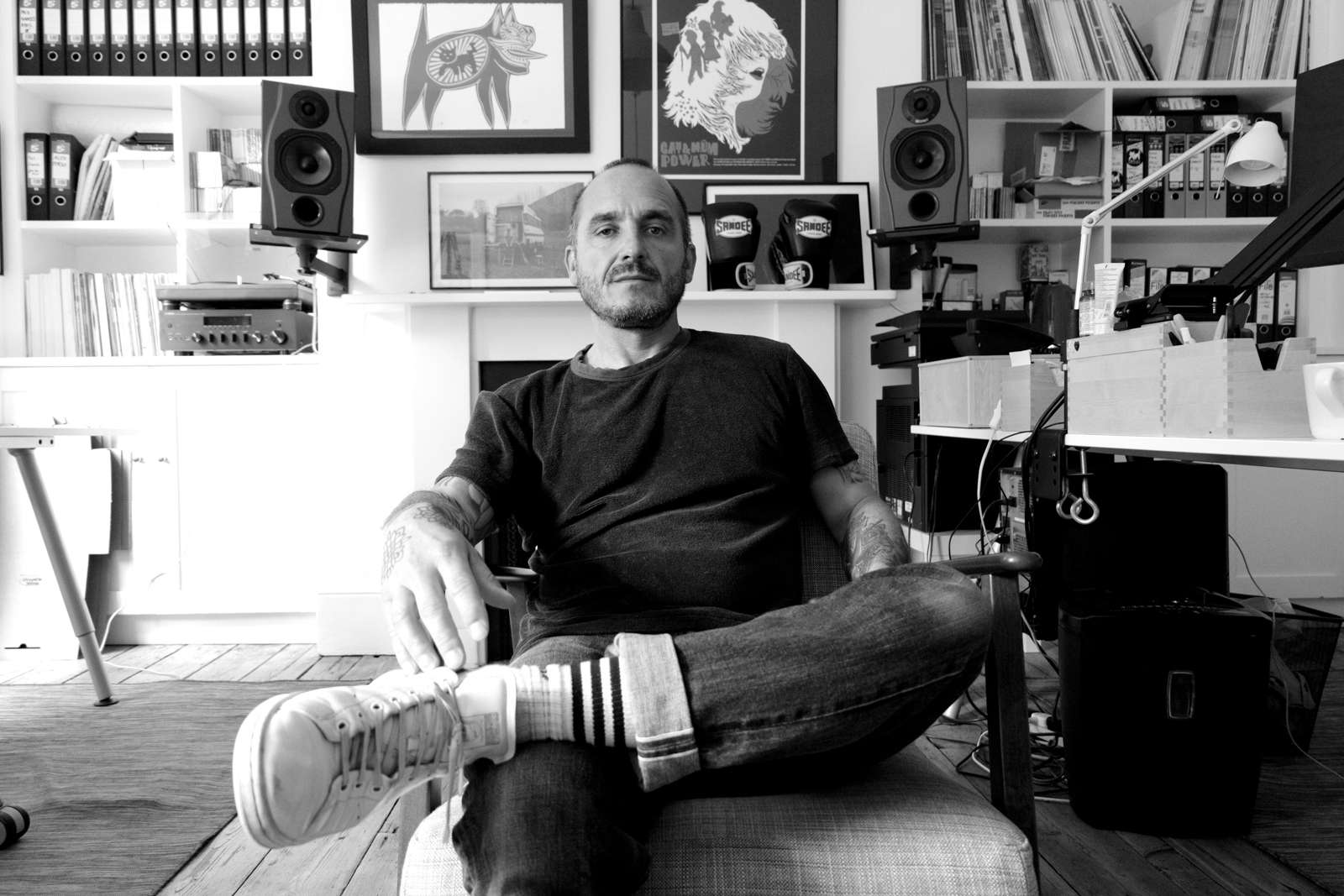 Black-and-white photo of the shop owner seated in a listening room, surrounded by shelves of vinyl, posters, and studio monitors.