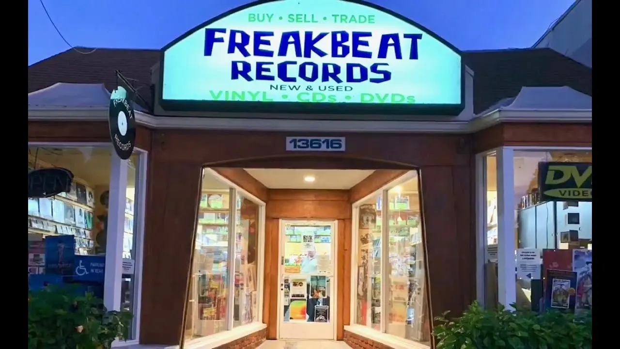 Front entrance of Freakbeat Records in Sherman Oaks, glowing under a bright blue evening sky. The store’s sign reads “Buy • Sell • Trade – Vinyl, CDs, DVDs,” with record-lined walls visible through large display windows.