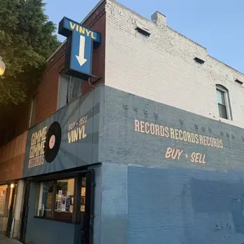 Street view of Gimme Gimme Records in Los Angeles, a corner shop with retro signage reading “Buy + Sell Vinyl” and a big blue arrow pointing to the entrance. The painted exterior gives it a vintage neighborhood vibe.