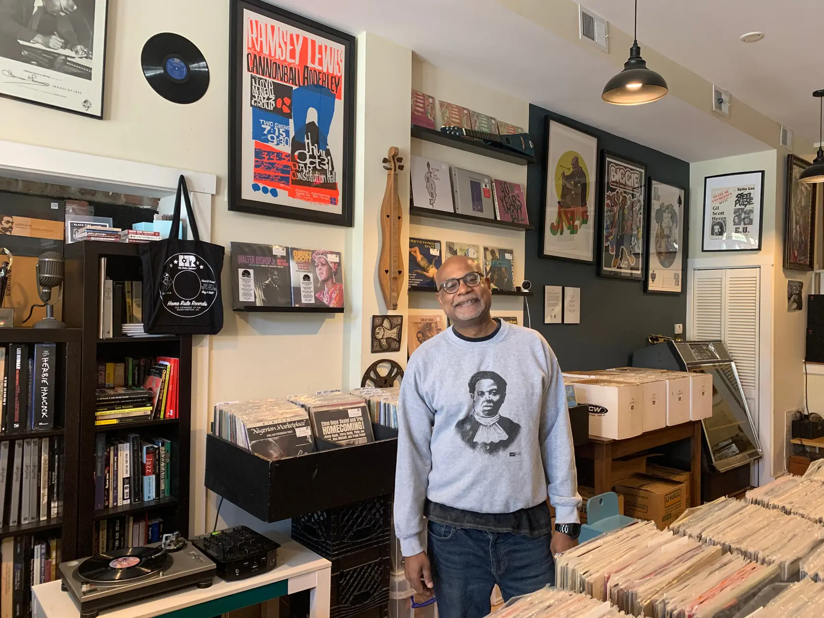 Owner standing among tightly packed bins in a cozy room, surrounded by framed jazz/hip-hop posters, books, and cassettes.