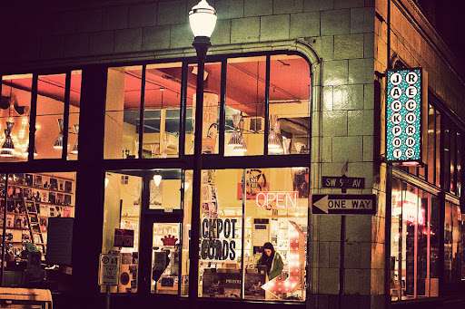 Nighttime view of Jackpot Records in Portland, Oregon, illuminated by warm lights from inside the shop. The neon “OPEN” and “Records” signs glow through the large windows, giving the storefront a nostalgic, welcoming vibe for late-night vinyl hunters.