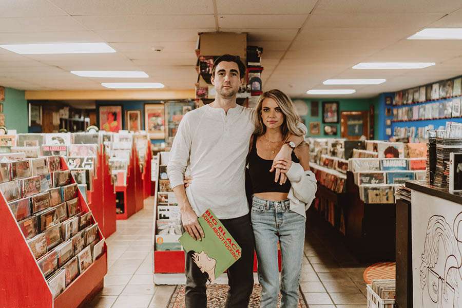 A couple standing in the middle of Long in the Tooth Records in Philadelphia, surrounded by rows of vinyl albums and red record bins, holding a Big Black LP.