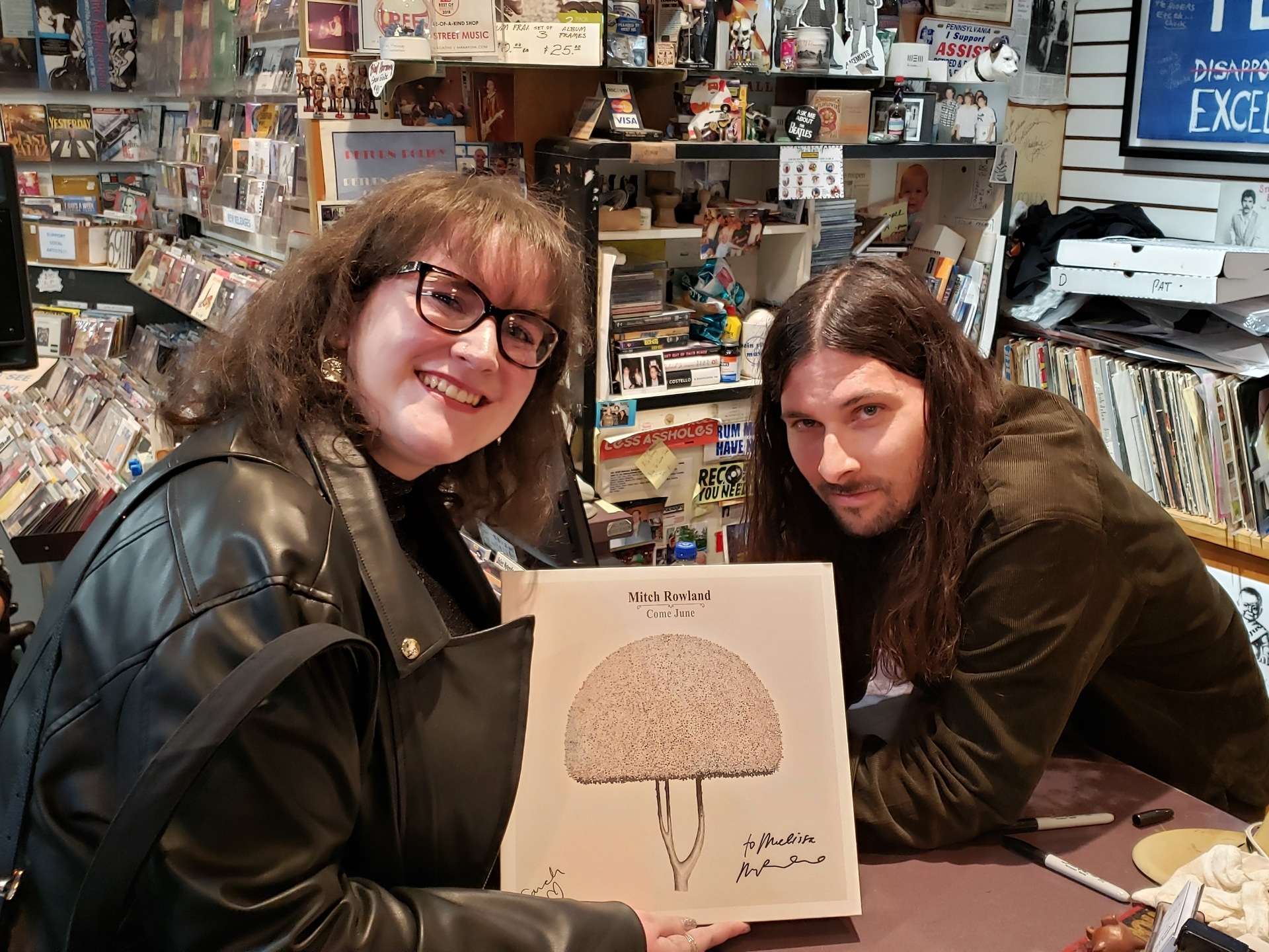 A smiling customer posing with musician Mitch Rowland inside Main Street Music in Philadelphia, surrounded by CDs, vinyl, and posters in a cozy shop interior.