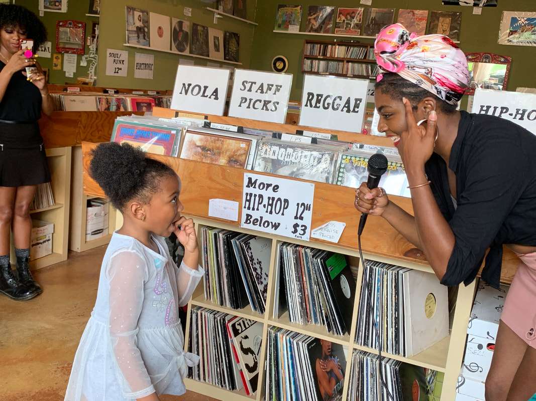 Inside NOLA Mix Records, a vibrant community record shop where a woman with a microphone talks to a young girl among crates labeled NOLA, Reggae, and Staff Picks.