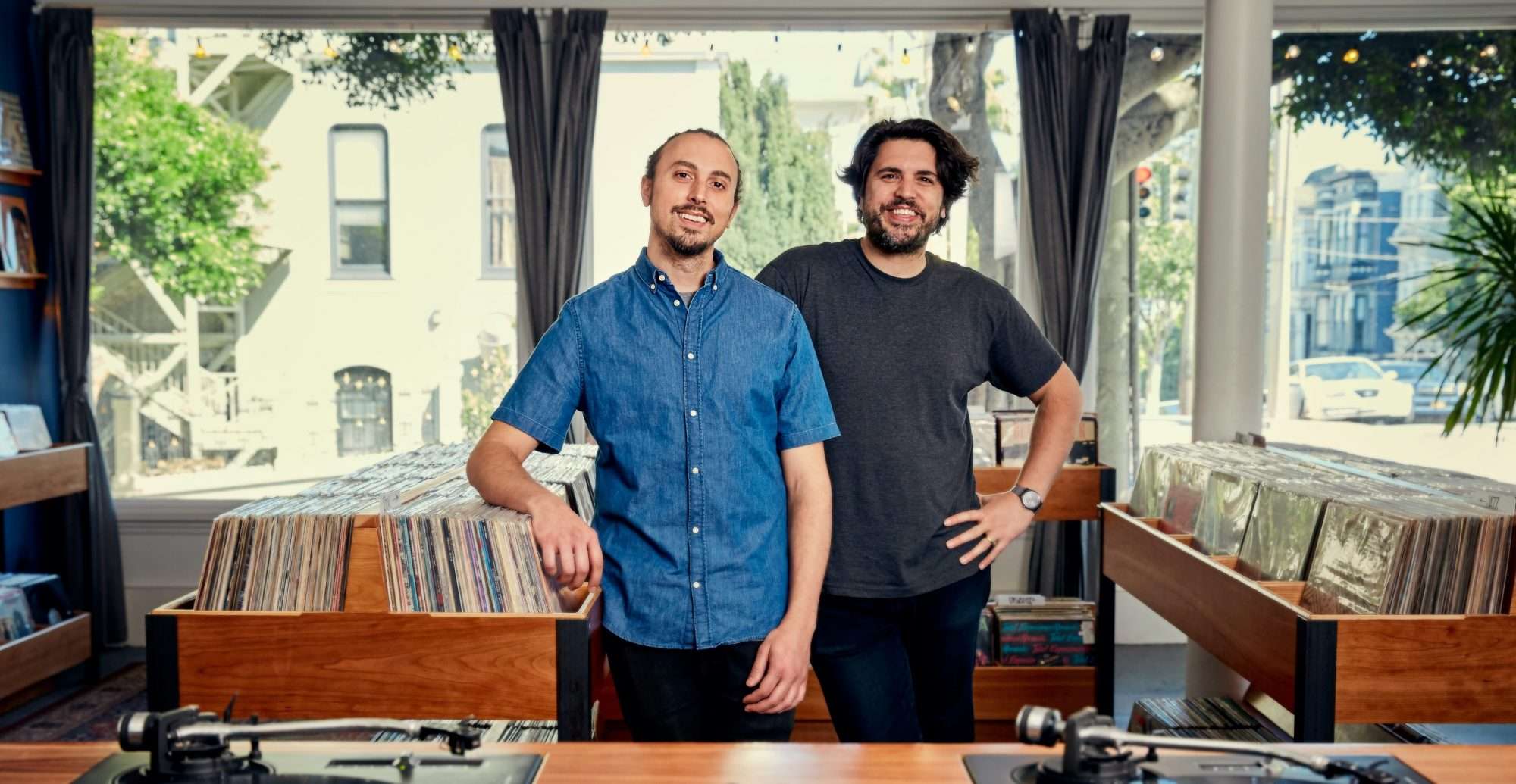 Two owners standing inside a bright Mission District record store with sunlight, large front windows, and rows of neatly organized vinyl bins.